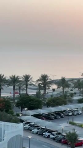 This video showcases a serene coastal landscape at sunrise or sunset, likely in Ras Al-Khaimah, United Arab Emirates. The sky is a soft gradient of pink and orange, indicating either the early morning or late evening. The calm, pale sea stretches to the horizon, reflecting the muted colors of the sky.

The foreground features a well-maintained parking lot filled with various cars, some covered by white, arched awnings. Palm trees are strategically planted throughout the scene, adding a tropical feel, and are interspersed with lush green bushes and trees. A modern building with a white facade and prominent signage with green and blue accents is visible amidst the palm trees, suggesting a commercial or recreational facility.

The overall atmosphere is peaceful and tranquil. The gentle lighting and the vastness of the sky and sea evoke a sense of calm and natural beauty. The presence of cars and the structured parking area indicate a developed area, likely a resort or a public space near the coast. The video captures the quiet beauty of a coastal environment as the day transitions.