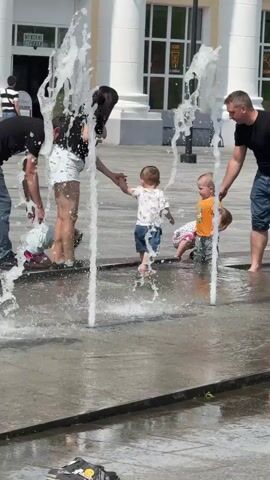 The video shows a group of people playing in a splash pad on a sunny day. Two adults, a man and a woman, are supervising three young children. The children are running around and playing in the water jets. The man is holding the hand of one child, while the woman is reaching out to another. The splash pad is made of concrete and has several water jets that spray water into the air. In the background, there is a building with white columns and large windows.