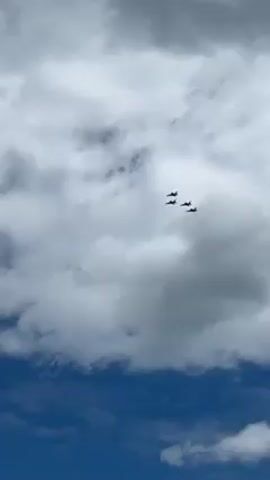 Certainly! Here's a detailed description of the video:

The video shows four fighter jets flying in formation against a sky that is partially blue with large, puffy white clouds. The camera pans down to reveal a group of people standing on a beach looking up at the jets. Some of them are wearing hats and casual clothing. The ocean is visible behind them. The overall atmosphere seems like an air show or a special aerial display.