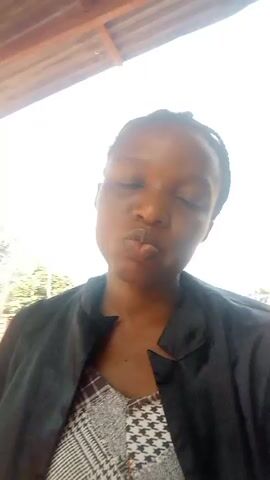 A woman with dark skin is taking a selfie. She is wearing a patterned top and a black jacket. Her hair is pulled back. Behind her is a person in a red shirt with white lettering. The background is bright and out of focus, but appears to be outdoors with some buildings visible. The woman smiles and touches her face.