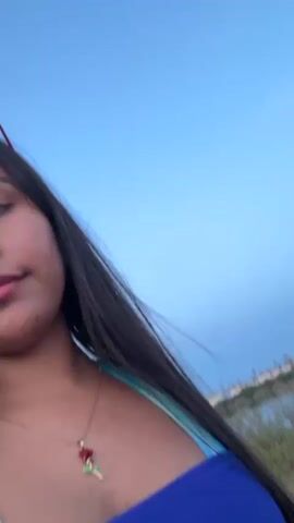 A woman with long dark hair wearing a blue and turquoise bikini and a necklace with a red rose pendant is walking outdoors near a body of water. She turns her head to the right and smiles at the camera. The sky is a clear blue and the sun is low in the sky, suggesting it is either sunrise or sunset. There are some bushes and trees in the background.