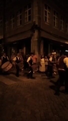 A street parade is taking place at night, featuring a group of men dressed in vests and white shirts, playing large bass drums. They are marching down a cobblestone street lined with buildings. The street is dimly lit by lamps mounted on the buildings. Some spectators can be seen on the sidewalks, including people wearing light-colored dresses. The parade appears to be moving forward, with the drummers in motion.