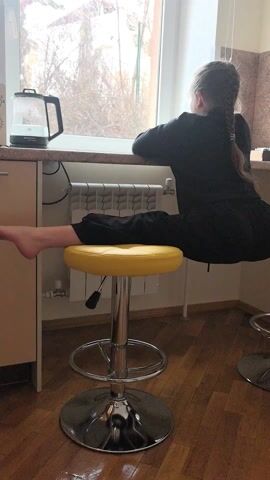 Here's a detailed description of the video:

A young girl with a long braid is doing a split between two yellow bar stools in a kitchen. She is facing a window and leaning on the countertop. She is wearing a black outfit.  The stools have chrome bases. Behind her are countertops and a window, as well as a radiator. A kettle is visible on the counter. The floor is wooden.
