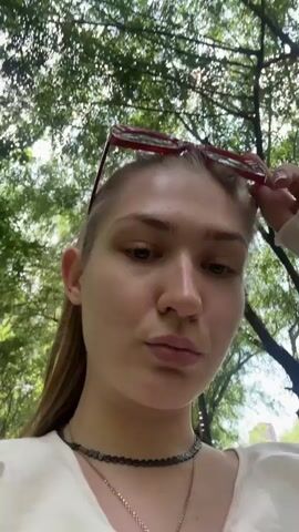 This video shows a young woman with blonde hair, wearing red-framed sunglasses and a white top, looking at the camera with her lips puckered. She is outdoors, with trees and foliage in the background, and sunlight filtering through the leaves. Another person, a male with dark hair wearing a grey shirt and a black baseball cap with a Red Bull logo, is visible behind her, looking at a phone. The camera appears to be handheld and is focused on the woman. The video is short and appears to be a selfie-style recording.