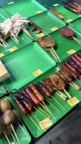 The video shows a variety of food items displayed on green banana leaves arranged on a market stall. The items are mostly on skewers and include sausages, potatoes, broccoli, cauliflower, mushrooms, eggs, and various types of meatballs and processed meats. Some of the items appear to be grilled or cooked. There are also small containers of sauce and packets of instant noodles visible. The stall seems to be a street food or market stall, with pricing labels visible on some of the items. The overall impression is of a diverse selection of ready-to-eat food options.