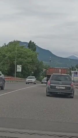 This is a vertical video shot from the perspective of a passenger in a vehicle driving on a multi-lane road. The road appears to be a highway or a major thoroughfare, with clear lane markings. In the foreground, a dark grey station wagon with a Russian license plate is prominently featured. Further ahead, several other vehicles are visible, including a lighter colored sedan, a pickup truck, and a large red truck. The road is flanked by trees and a bridge railing on the left, and on the right, there are signs and more trees. In the background, a range of mountains dominates the landscape under a cloudy sky, suggesting a potentially overcast or moody weather. The time of day appears to be daytime, given the ambient light.

There are some visible signs with text in Russian. One sign on the left, near a lamppost, has some text and a graphic that is difficult to read clearly. Another sign on the far right also displays Russian text.

The overall scene depicts a typical driving experience on a road in a mountainous region, likely in Russia given the license plate and text. The activity is straightforward: a vehicle is in motion, traversing the road. There are no discernible human figures within the frame, and the focus is on the road, the vehicles, and the natural scenery. The mood conveyed is neutral, simply documenting a journey.