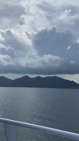 The video shows a seascape with a mountain range in the distance under a sky filled with large, grey and white cumulus clouds. The water is calm with a slight ripple and appears a dark blue-grey color. In the foreground, a white railing is visible, suggesting the view is taken from a boat or ship. The wind noise can be heard in the background.