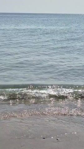The video shows a gentle wave breaking on a sandy beach. The water is calm with a few ripples, and the sand is a light gray color. In the distance, there are some rocks and a clear blue sky. The lighting suggests it is daytime, likely a sunny day. No people are clearly visible in the foreground, but there appear to be some figures further down the beach on the left side in the later frames.