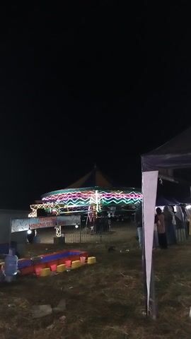 This is a video of a nighttime scene at an outdoor event, possibly a fair or festival. A large tent with colorful lights is the central focus, with people visible standing and walking around it. In the foreground, there is a small amusement ride, possibly a carousel or a similar attraction, with bright yellow and red seats. The ground is covered in dry grass, suggesting it might be an open field. The overall atmosphere appears festive and lively, with the bright lights contrasting with the dark night sky.

There is visible text on a sign that reads "PERMAINAN IKAN MEMANCING SESI". This likely translates to "Fish Game Fishing Session" or a similar description of a game. The time of day is clearly night, indicated by the dark sky and the illuminated structures. The presence of the tent and the amusement ride suggests an event is in progress. The weather appears clear as there are no indications of rain or strong winds. The location context of Sambirata, Indonesia, places this scene in a specific geographical area.