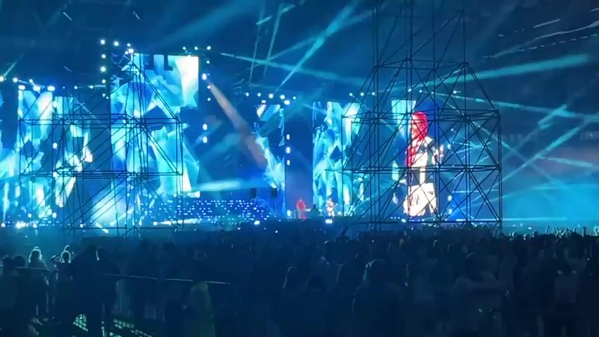 This is a video of a live concert. The stage is large and decorated with scaffolding and numerous screens displaying abstract blue geometric patterns. A performer, wearing a white outfit with black detailing and red hair, stands on the stage, facing the audience. A crowd of people can be seen in the foreground, watching the performance. The lighting is predominantly blue, with spotlights creating beams of light. The atmosphere appears energetic and visually stimulating.