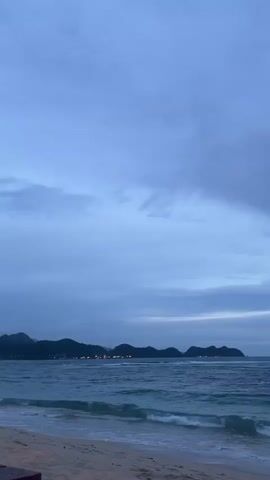 A video of a beach with waves crashing on the shore. The sky is overcast with clouds, and the water is a deep blue. In the distance, there are islands with hills. The video is in portrait mode and seems to be filmed at dusk or dawn.