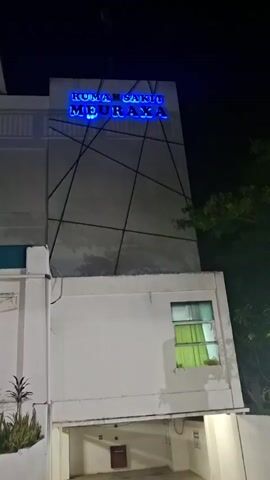 The video shows a nighttime exterior shot of a building at an angle. The building is primarily white and grey, with some green curtains visible through a window on the right side. A blue neon sign on the upper part of the building reads "RUMAH SAKIT MEURAXA". There are dark lines forming a geometric pattern on the grey section of the building. The overall lighting suggests it is dark outside.