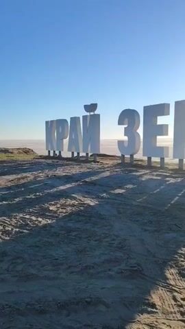 The video pans across a beach with large, white, three-dimensional letters spelling out "КРАЙ ЗЕМЛИ". The sun is visible in the background, shining brightly and creating long shadows from the letters. The sky is clear and blue. The ground is sandy with tire tracks visible. A small bird is perched on the top of one of the letters.