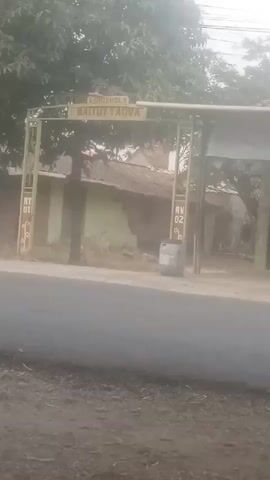 The video shows a street view with an archway entrance to a residential area. The archway has text that reads "Baitut Taqva" and "RTO1" and "RW02". A white car drives by on the street in front of the archway. The area appears to be rural or suburban with trees and simple buildings visible behind the archway. The lighting suggests it is daytime, possibly overcast.