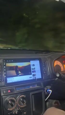 The video shows the interior of a car driving at night. The car's dashboard is visible, including a large touch screen playing a video. The steering wheel is partially visible on the right. The video is taken from the driver's perspective. The car appears to be on a road with streetlights and some sparse roadside vegetation.