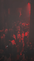 Nightclub crowd dances under red lights as patrons record scene
