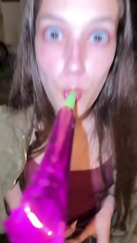 A person wearing a brown zippered hoodie lifts a glass with a red drink to their lips. Another person in a red tank top, blue jeans, and a light brown jacket holds a purple party horn to their mouth and smiles at the camera, then bends over to grab a glass with a red drink from the ground.