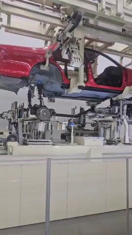 A red car body is suspended from an overhead conveyor system in a factory. Below the car body, a complex assembly of robotic arms and machinery is in operation. The machinery appears to be working on the underside of the car, potentially installing or inspecting components such as the suspension and exhaust system. The environment is a well-lit industrial setting with visible structural elements and support beams of the factory.