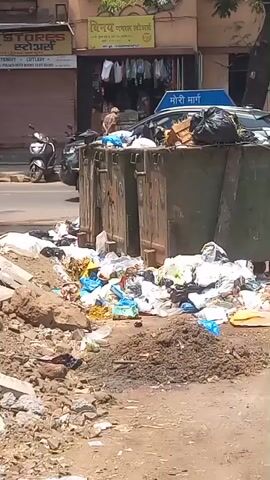 The video shows a street scene. First, an overflowing dumpster is visible, surrounded by trash. The camera then pans to the side, showing an excavation with exposed blue and yellow pipes on the ground.