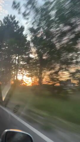 The video appears to be taken from inside a moving vehicle, looking out the side window. A road is visible in the lower portion of the frame. Trees line the side of the road, silhouetted against a sunset. The sky is a mix of blue and clouds, with orange and yellow hues from the setting sun peeking through the trees. A building can be seen in the distance. The camera is moving, causing the foreground to blur.