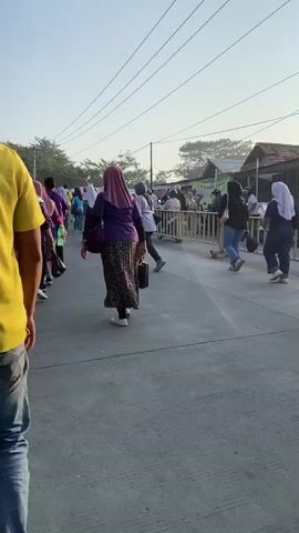 A group of people, mostly women wearing hijabs and purple shirts, are walking along a paved path. The women are walking in the same direction, away from the camera. Some of the women are carrying bags. In the background, there are trees and buildings. There is a metal fence along the right side of the path. The sky is clear and the sun is shining.