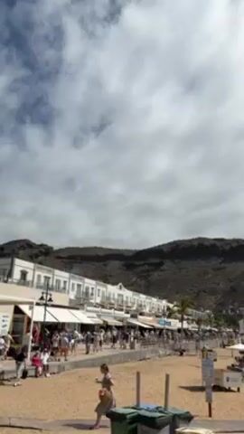 The video shows a beach scene, with a line of buildings and awnings along the edge of the beach. People are walking along the promenade in front of the buildings. On the sandy beach, many white umbrellas provide shade for sunbathers. The sky is partially cloudy, with patches of blue sky visible. In the background, a large, rocky hill rises behind the buildings. Two green garbage bins are in the foreground.