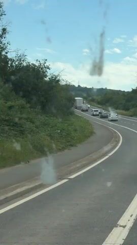 The video shows a highway scene in Long Ashton, United Kingdom, on a sunny day. The camera is positioned inside a vehicle, looking out onto the road. Cars and a large truck are visible on the highway, appearing to be in traffic or moving slowly. The road curves to the right, with a grassy embankment and trees lining the left side. The sky is blue with some clouds, indicating fair weather. The overall mood seems calm, possibly captured during a leisurely drive or while experiencing a minor traffic delay. There is no visible text in the video.