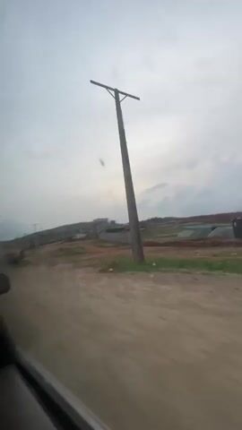 The video, shot from inside a moving vehicle, shows a view of a construction site on a cloudy day. The camera pans across a landscape of dirt and construction materials.  Several large concrete pipes lie on the ground near the roadside. A partially constructed building can be seen in the distance, enclosed by what appears to be a low wall. Power lines and utility poles line the perimeter of the site. The sky is overcast with grey and white clouds, suggesting possible rain. The road is unpaved and dusty.