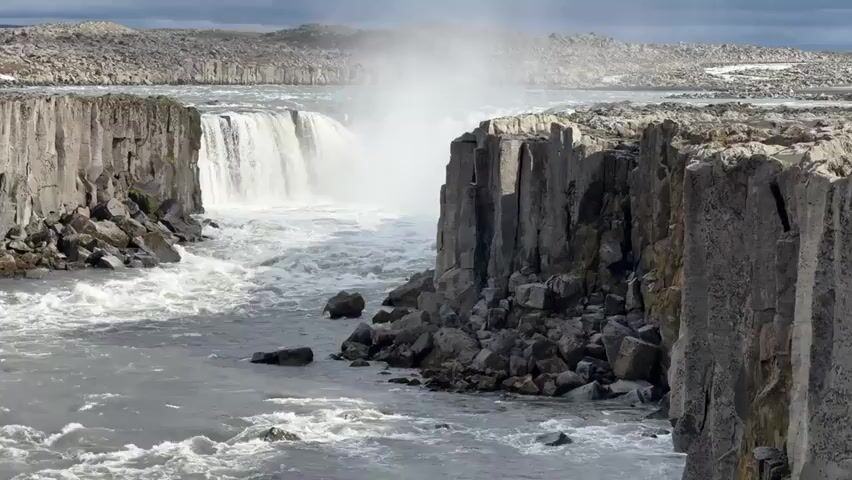The video captures a majestic waterfall in a rugged, natural setting, likely in Iceland. The powerful cascade of water, wide and forceful, plunges into a turbulent river. Steep, dark grey rock formations, resembling columnar basalt, frame both sides of the waterfall and the river as it flows away. Numerous large, dark rocks are scattered along the riverbanks and partially submerged in the churning water.

The river itself is a frothy, fast-moving expanse of grey-white water, exhibiting strong currents and many whitecaps as it flows through the gorge. A significant amount of mist rises from the base of the waterfall, indicating its immense power. The sky above is partially cloudy, casting a diffused light over the scene. No people or visible text are present in the video, emphasizing the raw, untamed beauty of the landscape. The overall impression is one of natural grandeur and immense geological force.