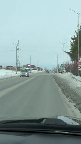 The view from inside a car moving down a paved road shows a typical winter scene in a rural or suburban area. The sky is overcast and the surroundings are generally flat, with snow piled along the sides of the road. A power transmission tower stands to the left of the road. Streetlights are spaced along the road, and various pedestrian crossing signs mark the street at intervals. A silver car passes in the opposite direction. In the background, smoke rises from a distant chimney. A building identified as a "shop" is visible on the right side of the road, marked by a sign in Russian. The overall impression is of a quiet, possibly cold, day on a relatively empty road.