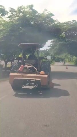 The video shows a tractor driving down a paved road. The tractor is orange and appears to be older. A person is driving the tractor. There are trees and power lines on either side of the road. The sky is bright and sunny. There is some dust or smoke coming from the back of the tractor. In the distance, there is a traffic cone and another person walking. The road appears to be in a rural or suburban area. The video is shaky, suggesting it was filmed from a moving vehicle.