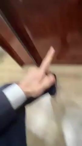 A blurred video shows a person sitting down, wearing a blue suit with a white cuff visible on the sleeve. They are holding a white bag. They extend their middle finger towards the camera. The background includes a dark wooden surface and a light-colored floor. The overall video quality is shaky and out of focus.
