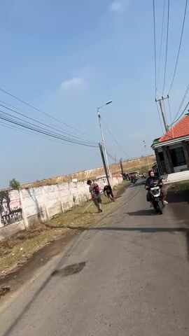 The video shows a street scene with several people and vehicles. On the left, there is a concrete wall with graffiti on it. Two people are standing near the wall, and one appears to be holding a long pole. A street light is positioned behind them. In the background, there is a dirt embankment and more graffiti on the wall.

The street itself is paved and appears to be a road. A motorbike with two people on it is riding away from the camera. Further down the road, more motorbikes can be seen. On the right side of the road, there is a building with a red-tiled roof and a small green structure. Overhead, there are power lines stretching across the scene. The sky is clear and blue.