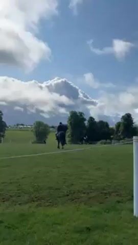 Here is a detailed description of the video:

The video shows a scenic outdoor equestrian event on a bright day with a blue sky punctuated by large, puffy white clouds. Two riders are seen on horseback in a large, green field. The first rider, dressed in a dark riding jacket and helmet, is closer to the camera and riding away from the viewer. The second rider is farther away and slightly to the right. They are also riding a horse, but details are less clear due to the distance. In the background, a line of trees frames the field, and in the far distance, what appears to be a gathering of tents or structures, possibly related to the event, can be seen. A white fence line is visible marking the edge of the riding area. The focus remains primarily on the riders and the expansive green landscape, creating a sense of open space and outdoor activity.