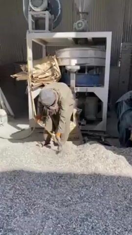 In a brightly lit indoor industrial setting, two individuals are engaged in manual labor around a large milling machine. The machine is elevated on a white frame and has several cylindrical components. A large fan is mounted on top. The first individual, wearing a head covering and gloves, uses a shovel to gather light gray shredded material from a sizable pile on the floor. He is focused on scooping the material and placing it into what appears to be a collection point on the milling machine. The second individual, also wearing a head covering and gloves, is closer to the machine, using a similar tool to handle the same material. There are partially filled burlap sacks next to the machine. The environment has corrugated metal walls, suggesting a warehouse or factory setting.