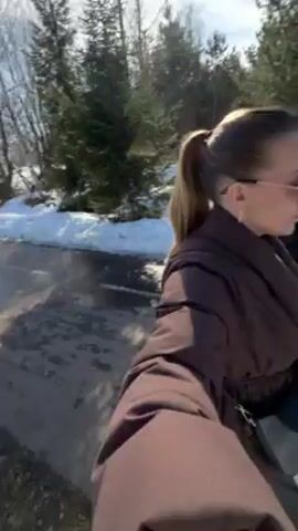 A family consisting of a man, woman, and baby is walking on a paved path on a sunny day. The woman holds the camera, showing herself in a brown puffer jacket and sunglasses. Her hair is pulled back in a ponytail. To her left, a man is pulling a black bag. Next to him is a baby sitting in a stroller. The baby is wearing a white outfit and hat. The ground is mostly paved but there are patches of snow on the side of the path. There are trees in the background under a bright blue sky.