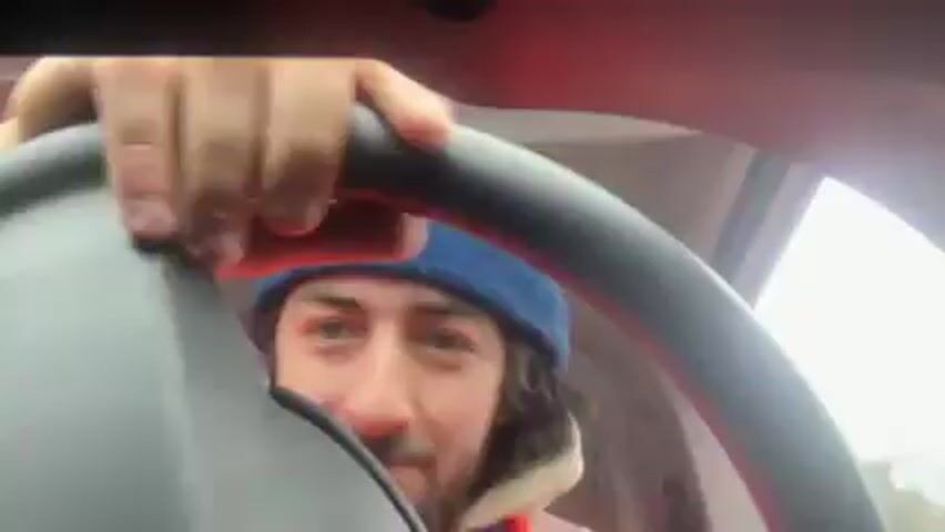 From the driver's seat perspective, a man wearing a blue beanie and orange jacket is shown operating the steering wheel of a vehicle. The view is somewhat obstructed by the steering wheel itself and the dashboard. The man looks directly at the camera, smiling and seeming cheerful as he drives. The background is visible through the windshield but is indistinct, suggesting movement or out-of-focus scenery. The light appears to be diffused, suggesting overcast conditions.