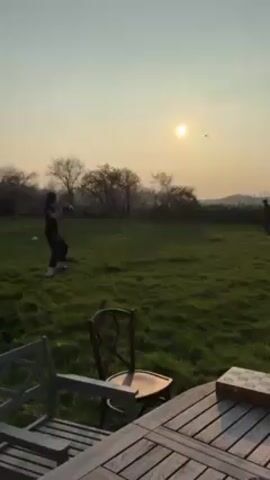 Two people are throwing a frisbee in a grassy field. The sun is setting in the background. In the foreground, there is a wooden table and chairs on a deck. A decorative box is on the table. One person is wearing a dark shirt with a number on the back. The other person is standing further away in the distance.