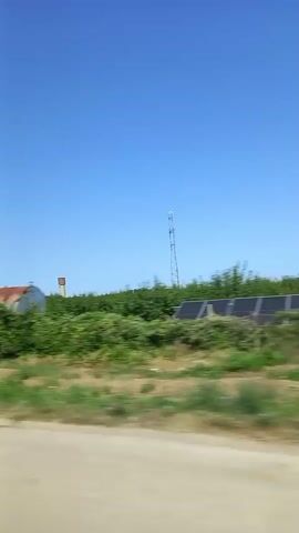 The video shows a landscape view from a moving vehicle. The sky is a clear blue. In the foreground, there is a dirt road and green vegetation. In the middle ground, there are solar panels and trees, as well as a large, rusty cylindrical tank with a tower structure on top. Further in the distance, there is a vast field of sunflowers, and a water tower. The overall scene depicts a rural or agricultural area on a sunny day.