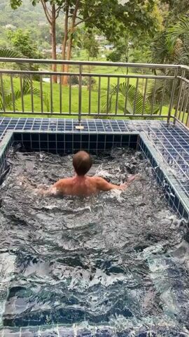 A person is partially submerged in a rectangular hot tub with blue tiled edges. The person is facing away from the camera, with their head turned to the side, and appears to be moving their arms in the water, creating splashes. The hot tub is situated on a tiled patio, with a metal railing in the background. Beyond the railing, a lush green landscape of trees and hills is visible under a cloudy sky.
