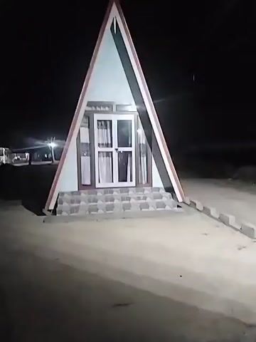 The video shows a triangular-shaped house at night. The house has a white and light blue exterior with a brown trim. There is a white door in the center, with two white curtains visible through the glass. There are also a few steps leading up to the door, made of grey tiles. The surrounding ground appears to be sandy. The background is completely dark, except for a few lights in the distance to the left. The camera remains mostly static during the video.