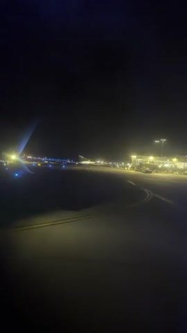 The video shows an airport tarmac at night. The perspective is from an airplane window, with the wingtip visible on the left side of the frame. The tarmac is lit by blue runway lights and yellow floodlights from the terminal buildings. Several other airplanes and airport vehicles are visible in the distance. The overall scene is dark, with the lights of the airport providing the only illumination.