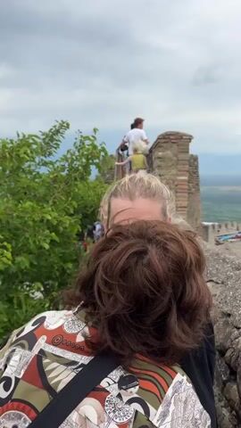 The video shows a group of people climbing a stone staircase on the exterior of a historic structure, possibly a castle or fortress. A lush green tree with red berries is prominently featured in the foreground, partially obscuring the view of the people. The sky is overcast with dark clouds. The people are dressed in casual summer clothing. They are ascending the stairs in a single file line, with some carrying bags. The stone walls of the structure are made of irregular, rough-hewn stones, and there is a brick parapet at the top. In the background, a scenic landscape with rolling hills and some buildings can be seen under the cloudy sky. The camera perspective is from behind the group, looking up the stairs.