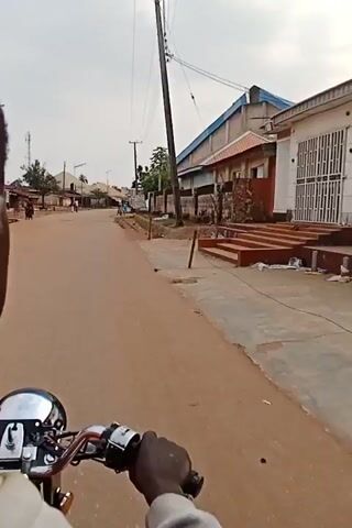 The video shows a point-of-view shot from someone riding a motorcycle down a street in what appears to be a rural or developing area. The road is mostly dirt with some paved sections. Buildings line the street, including some with corrugated metal roofs and others with more substantial construction. There are power lines overhead. A few people are visible on the street, some walking and others on motorcycles. The sky is overcast. The camera is focused on the road ahead and the motorcycle's handlebars.