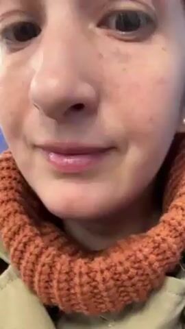 A close-up shot shows a woman applying lip gloss with a wand applicator to her lips. She wears an orange knitted collar.  She has fair skin with some freckles visible. Her eyes are brown.  She is wearing a tan or beige jacket. The background is slightly blurred, showing the interior of a train.