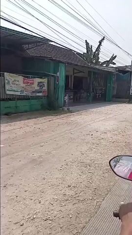 A dirt road passes by a row of shops or market stalls. The buildings have open fronts, some with awnings and displays of goods. On the right side of the road, a motorcycle is visible, suggesting the video is taken from the perspective of the rider. There are a few people in the scene, including a woman and child near one of the stalls. The environment appears to be a rural or semi-urban area, with overhead power lines and trees visible.