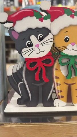 The video shows a close-up shot of several wooden cat decorations, each wearing a Santa hat and a scarf. The cats are of various colors, including black, orange tabby, and gray. The decorations are arranged side-by-side on a surface, likely a shelf or display. The background is blurred, but it appears to be an indoor setting, possibly a store or craft fair, with shelves of other items visible. The lighting is bright and appears to be artificial. The camera pans slowly across the decorations, focusing on their details and festive appearance.