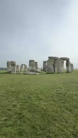The video presents a wide shot of Stonehenge, set against a backdrop of a grey, overcast sky and sprawling green fields. The ancient stone structure, composed of large, grey stones arranged in a circular pattern, dominates the scene. The grass in the foreground is short and well-maintained. In the distance, small figures of people are visible, but they are too far away to discern specific details. There is minimal movement in the video, and the camera remains static, offering a fixed view of the landscape and the historical monument.