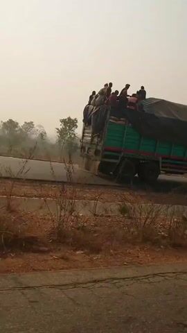 A medium shot reveals a green truck with a black tarp driving on a paved road. The bed of the truck is covered with the tarp and several people are seated on top of it. Dry, brown foliage is visible on the roadside. The sky is hazy. The camera follows the truck briefly as it drives past. The view then becomes blurry as the camera seems to lose focus.