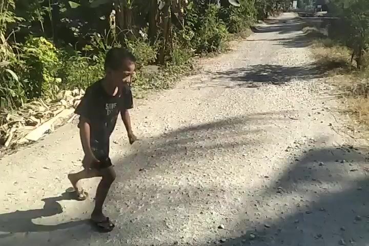 A young boy in a black t-shirt and shorts runs along a dirt path next to a canal. He stops at the edge of the canal and reaches towards something in the water. The boy is on the right side of the path and the canal is on the right side of the path. The path is surrounded by vegetation and trees. The sun is shining and casting shadows.